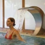 donna in una piscina termale che si vede a mezzo busto sotto una piccola cascata di acqua termale